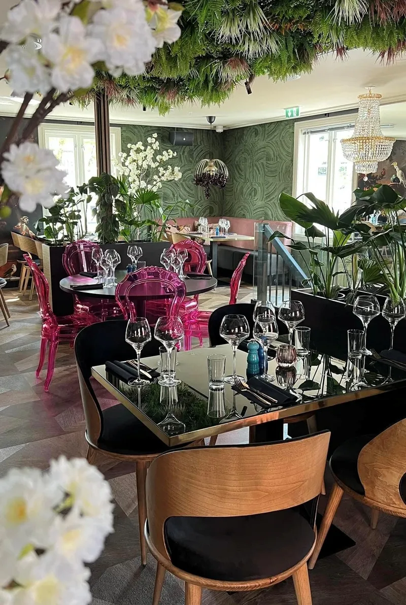 Restaurant Alma – Tønsberg: spisesal med blomster i taket, rosa stoler, dekkede bord og en krystallkrone foran grønne vegger