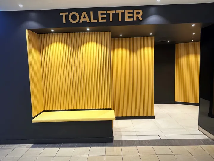 Publikumstoaletter – Alti Farmandstredet: inngangsparti til toalettene med gul lamellvegg, gul benk og skiltet «Toaletter» på en mørkeblå vegg