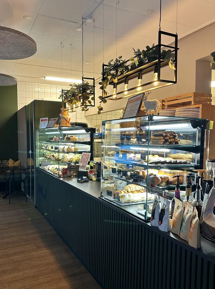 Den glade baker – Asker: bakeridisk med glassmontre fulle av bakevarer, mørkt lamellpanel og planter i hengende hyller over