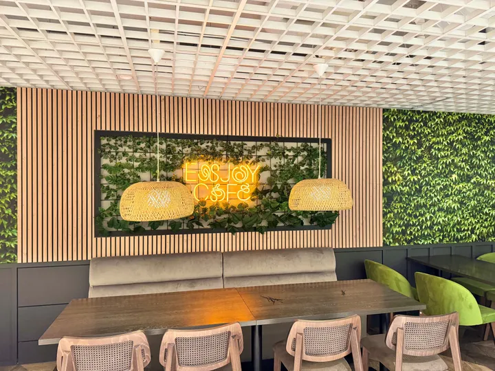 Enjoy Cafe – Hammerfest: langbord med flettede stoler foran trelameller og et felt med grønne planter og neonskiltet «Enjoy Cafe» mellom to kurvlamper
