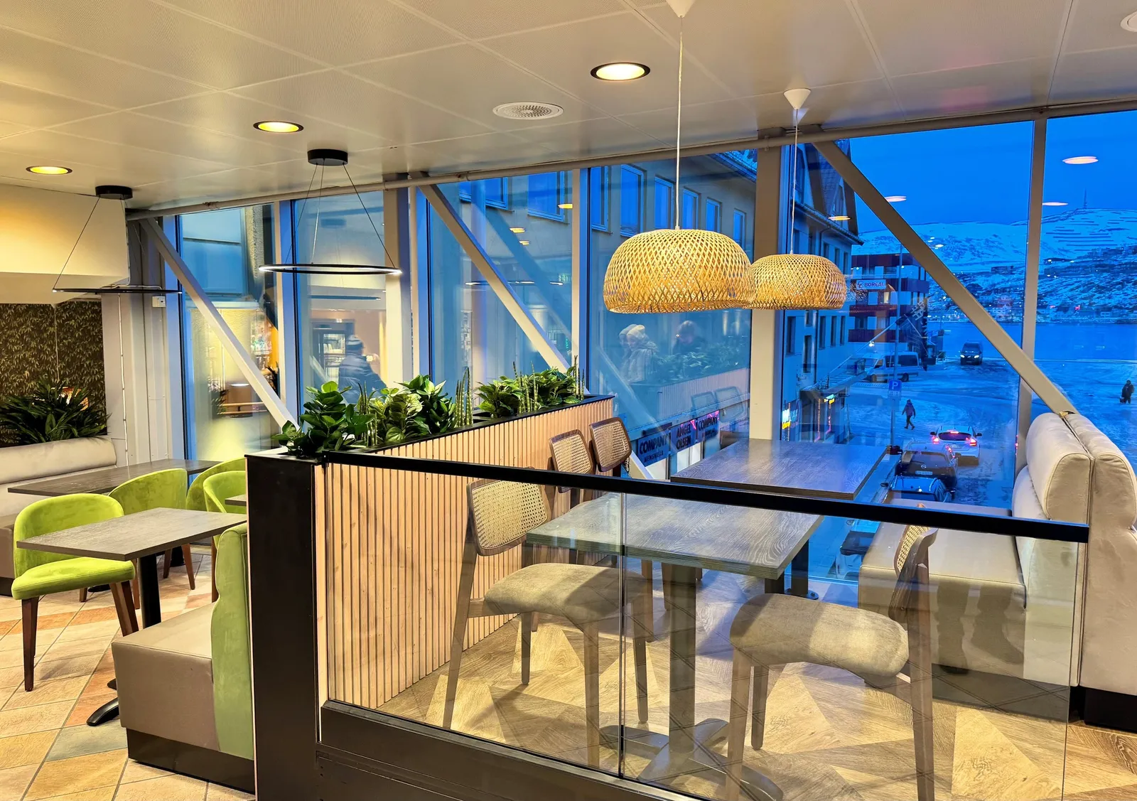 Enjoy Cafe – Hammerfest: sittegruppe med limegrønne stoler og kurvlamper ved store vinduer med utsikt over havnen i blått lys