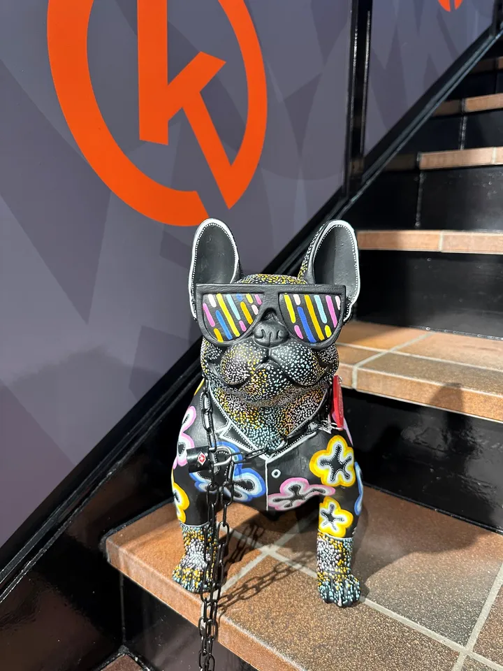 G Hagen-Norli – Hammerfest: fargerik bulldog-figur med solbriller ved en trapp, foran en grå vegg med en stor oransje logo